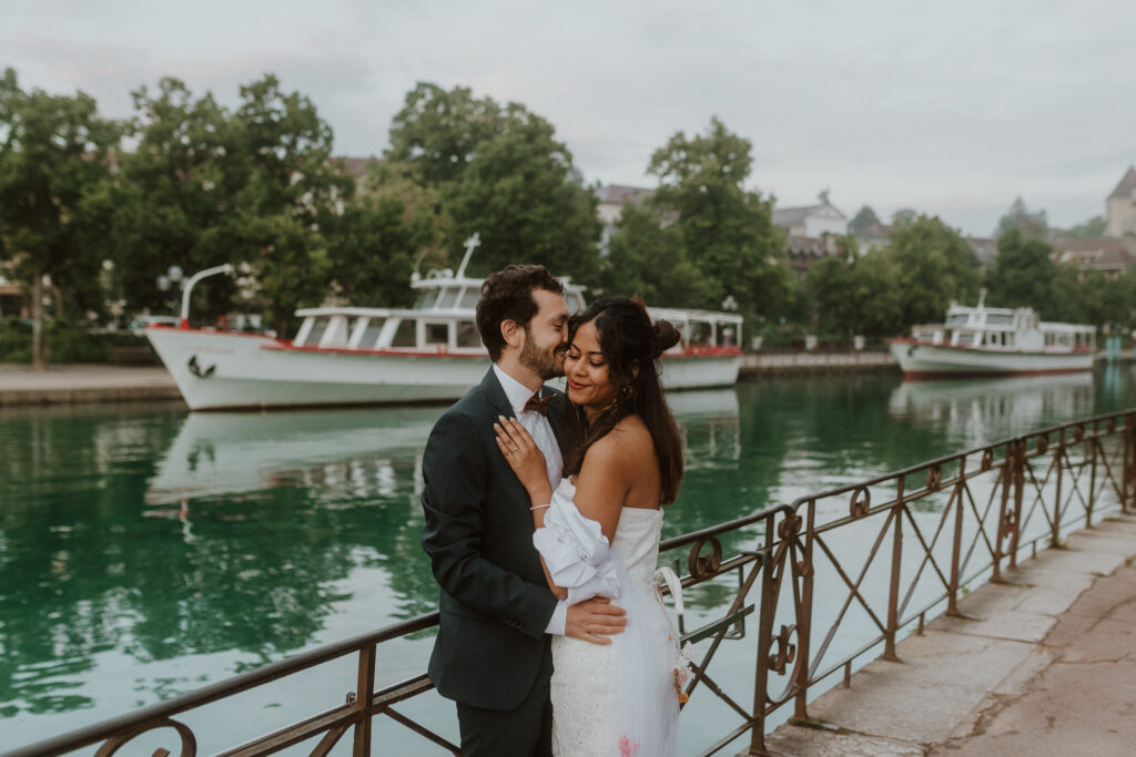 Photo d'un couple en tenue de mariage devant le gâteau libellule à Annecy. L'eau est turquoise. La photo est prise par Chloé Laydevant au levé du soleil.